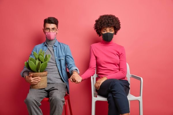 infected married woman and man have corona virus wear protective masks and hold hands sit on chairs and stay at home during self isolation or pandemic